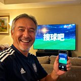 一位中年男性球迷在客厅观看比赛时的自拍，身后电视屏幕显示着搜球吧的直播画面